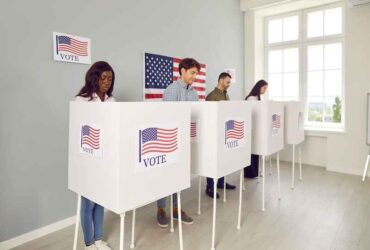 Portrait of a group of young happy diverse American citizens people men and women standing at vote center with USA flags in voting booth putting their ballots in bin on election day.