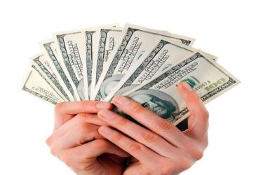 The image shows hands holding a fan-shaped arrangement of 100 US dollar bills.